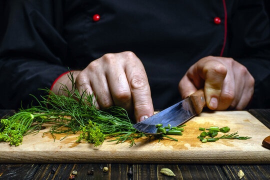 Professional cook finely chops green herbs on cutting board, using knife for careful prep. Hands guide dill and chive stems into small pieces, showing focused culinary concentration.