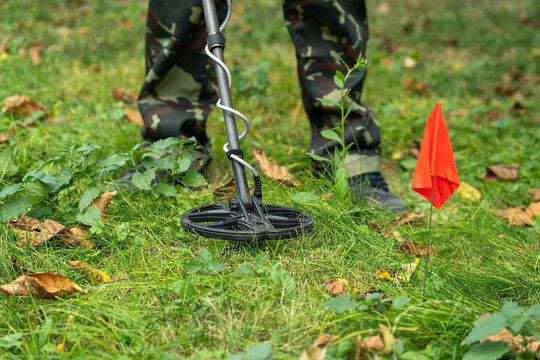 In the heart of a vibrant green field, a person clad in camouflage stands with a metal detector, searching for buried treasures among fallen leaves