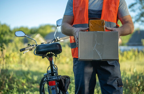 A courier in an orange vest stands in a lush, green field holding a box. A bicycle rests nearby, illuminated by bright sunlight on a vibrant day