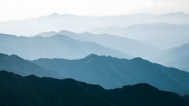 Peaceful landscape featuring layers of mountain ridges fading into a soft, hazy blue distance under a bright, overcast sky above