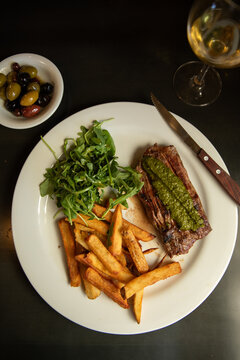 overhead shot of steak frite with chimichurri, fries, and arugula salad on a white plate with bowl of mixed olives, white wine, and a wood knife