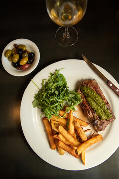 overhead shot of steak frite with chimichurri, fries, and arugula salad on a white plate with bowl of mixed olives, white wine, and a wood knife