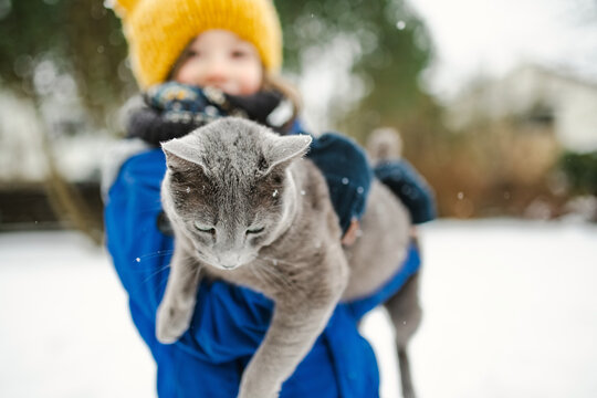 Young child holding a Russian Blue cat outdoors on a snowy winter day. Child and beloved family pet spend time together in cold seasonal weather.