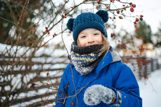 Happy young child playing with fresh snow in the backyard on a winter day. Smiling kid holding snow in gloved hands and enjoying outdoor seasonal activity.
