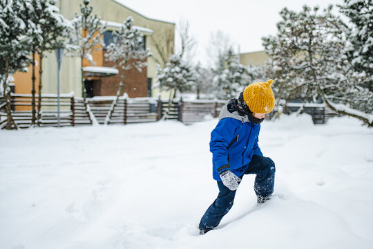 Happy young child playing with fresh snow in the backyard on a winter day. Smiling kid holding snow in gloved hands and enjoying outdoor seasonal activity.