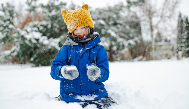 Happy young child playing with fresh snow in the backyard on a winter day. Smiling kid holding snow in gloved hands and enjoying outdoor seasonal activity.