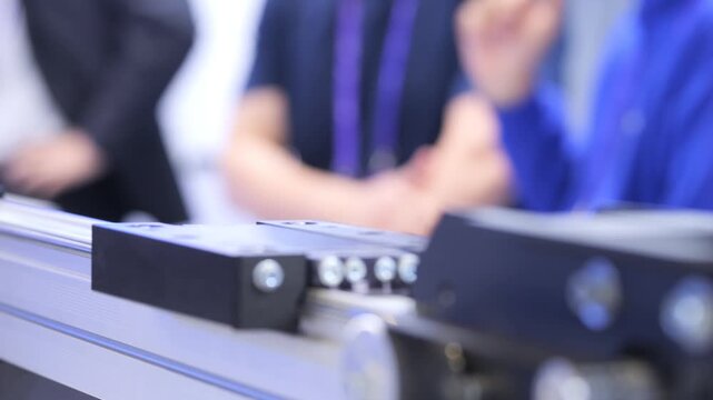 Close-up of industrial automation equipment on display. Blurred people in background at trade show. Focus on mechanical components and precision tools
