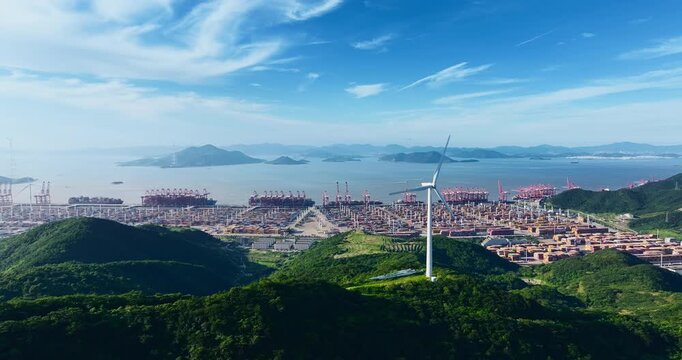 Aerial view of a coastal port terminal with shipping containers, heavy cranes, green hills, and a wind turbine under a clear blue sky.