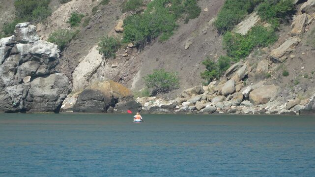 Kayak enthusiast paddles across a calm coastal sea near a rocky mountain cliff base during summer. Static long shot.