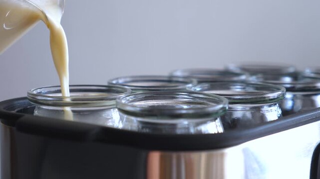Cream pours into glass jars in stainless steel caddy, filling containers for homemade preserves. Slow filling action continues with smooth beige liquid against clean kitchen backdrop, calm