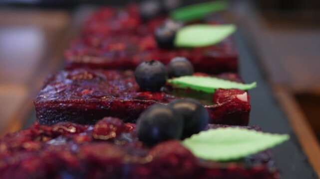 Blueberry topped cake bars sit neatly on black tray, their glossy berry surface and mint garnish creating elegant dessert presentation. Shallow focus adds warm bakery atmosphere and inviting feel.