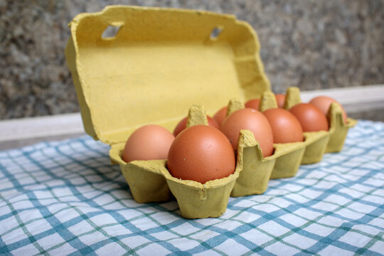 Open yellow egg carton filled with fresh brown eggs on blue checkered tablecloth, granite countertop background, farm fresh produce closeup shot, high quality stock photo