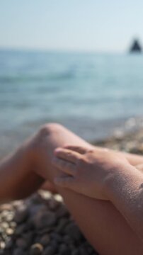 Cosmetic body care lotion applied to smooth legs by relaxed person sitting on sunny pebble beach. Vertical closeup shot.