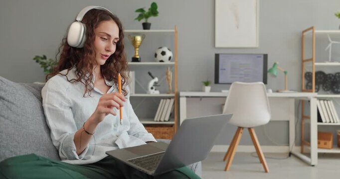 Woman student studies online with laptop headphones. Sitting on floor in home office, she takes notes with a pencil during remote lesson. Ideal for education, learning and study lifestyle.