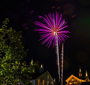 Colorful fireworks burst above buildings in a town at night. Trees surround the area, adding to the festive atmosphere. People watch the display from various spots.