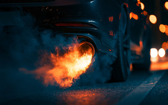 Close-up view shows car exhaust emitting flame and smoke during revving. The setting is at night on a street, highlighting the performance of the vehicle and its exhaust system