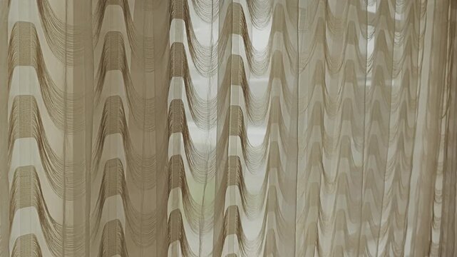 Delicate brown sheer string curtains with a wavy pattern gently moving in the breeze. Sun shining through the window creating soft light