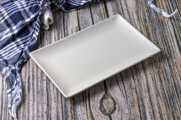 Empty white ceramic plate on rustic wooden background with glass and checkered napkin, top view...