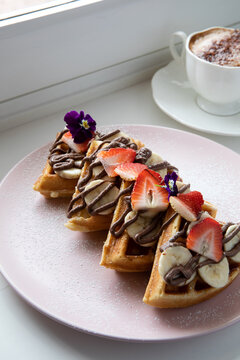 Plate of banana, strawberry and hazelnut cream waffle on a light pink plate, restaurant setting.