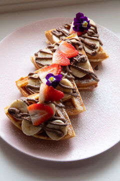 Close up of Plate of banana, strawberry and hazelnut cream waffle on a light pink plate, restaurant setting. 