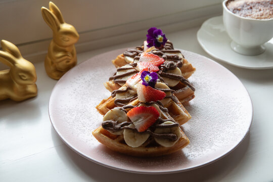 Plate of banana, strawberry and hazelnut cream waffle on a light pink plate, restaurant setting.