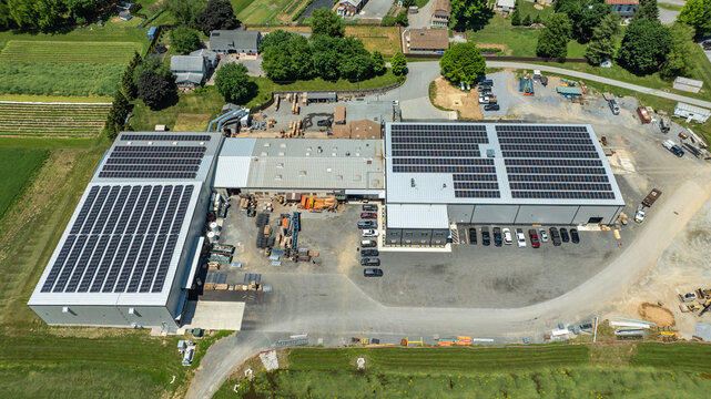 The view shows a large building with solar panels on the roof. Trucks and equipment are visible nearby. The scene is set in a rural area with green fields and trees surrounding the site.