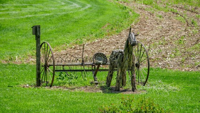 An old piece of farm equipment sits on green grass near a tilled land. The scene captures the blending of nature with remnants of farming life.
