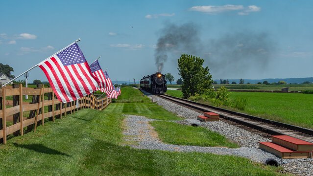 A steam train moves along the tracks near a green field. American flags line a wooden fence, fluttering in the wind. The scene takes place on a sunny day in a rural area.
