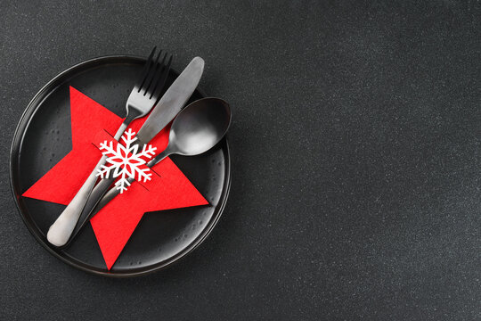 Christmas table setting with black plate and cutlery arranged in red star shaped cutlery holder decorated with white snowflake. Dark textured background with copy space, elegant holiday backdrop.