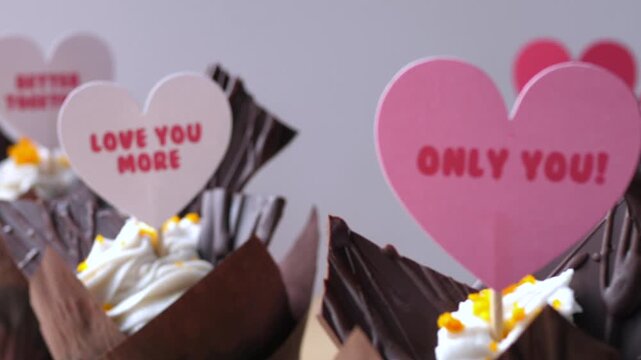Romantic chocolate cupcakes with heart decorations stand ready for gifting, each topped with cream and paper hearts. Only You and Love You More signs give dessert display tender celebratory feel.