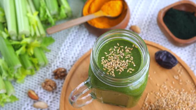Green smoothie in glass mug forms centerpiece among celery stalks, pumpkin, dates, walnuts and scattered seeds. Fresh blended beverage rests on wooden board, suggesting healthy nutrition and vitality.