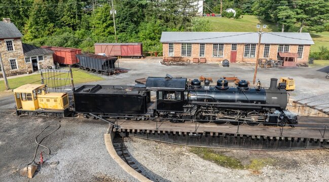 A steam train is located on a turntable in a railway yard. The setting includes several buildings and rail cars. The scene occurs in a rural area with trees in the background.