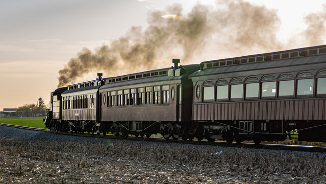 A steam train moves along tracks in an open field as the sun sets in the background. Smoke rises from the engine as the train approaches, creating a nostalgic scene.