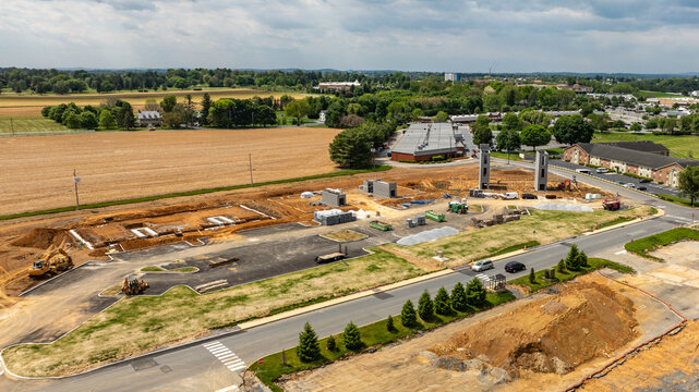 Workers are present at a construction site where new buildings are being erected. The location has open fields and some trees in the background. Machinery and materials are visible on the ground.