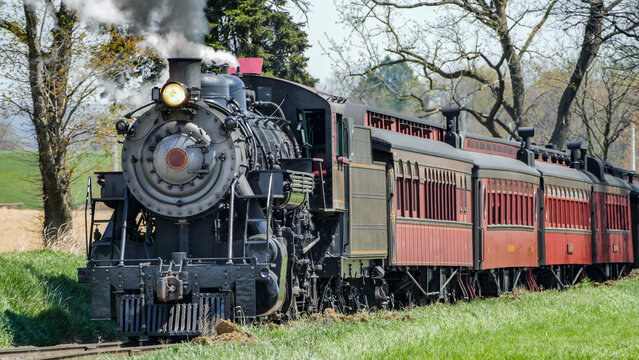 A steam train is traveling on tracks through a rural landscape. The train has several carriages and smoke is rising from the engine. There are trees and green grass close to the tracks.