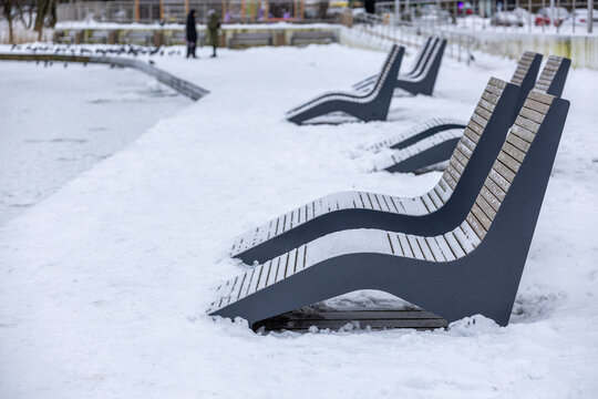 Snow-covered lounge chairs beside frozen pond in public city park on winter day