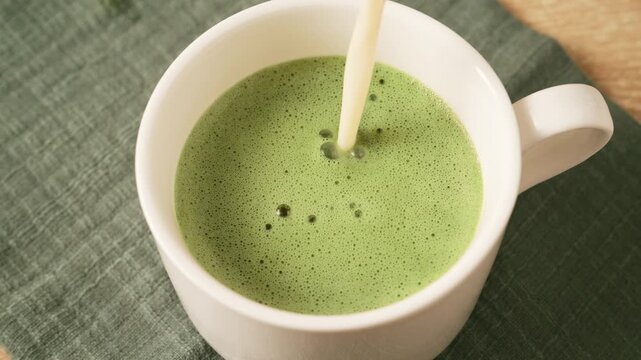 Pouring stream of oat plant milk into frothy green matcha tea in white ceramic mug. Antioxidant detox vegan matcha beverage for daily wellness routine.