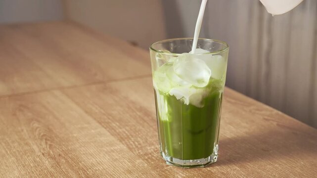 Pouring fresh milk over ice cubes into green matcha tea in tall glass. Preparing refreshing iced matcha green tea latte drink.