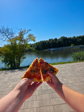 First person view of parent and kid hands holding two slices of tasty pizza outdoors in a sunny park with a scenic lake and green trees in the background.