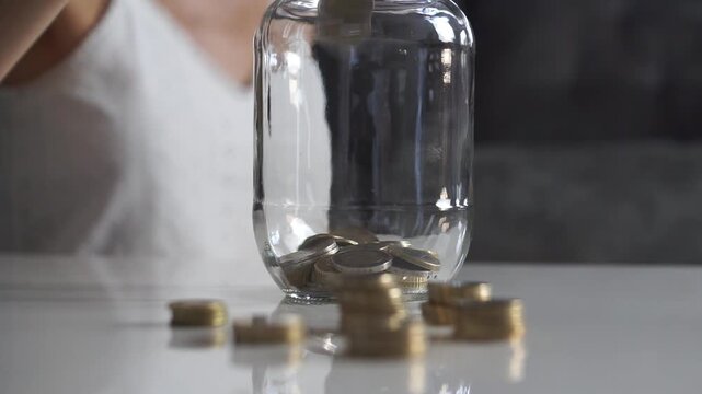 Low-angle close-up of euro coins being dropped into a clear glass jar on a white table with small stacks of coins in the foreground.