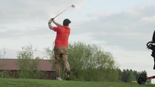 Male golfer in orange shirt prepares to swing a golf club on a lush green course, with trees and a barn visible in the background, showcasing the action of a golf shot