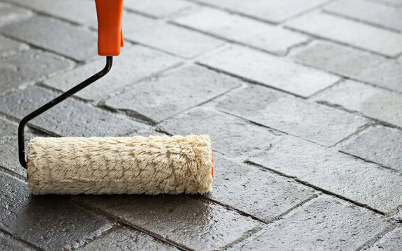 Construction worker applying gray cement-based coating with paint roller on paved surface