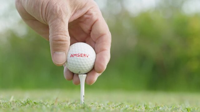 Close-up of hand placing a golf ball on a tee in lush green grass, with blurred background of trees, showcasing the preparation for a golf swing in a serene outdoor setting
