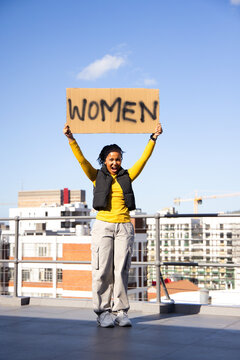 African woman holding cardboard WOMEN sign, shouting on rooftop in yellow top and black vest