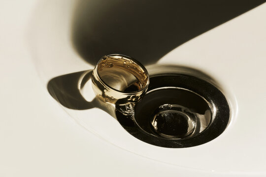 Single wedding band drain conceptual still life on white porcelain bathroom sink featuring unadorned gold ring balanced beside dark drain hole with crisp natural light and sharp shadow
