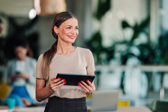 Professional businesswoman holding tablet, smiling and looking away, standing in modern coworking office