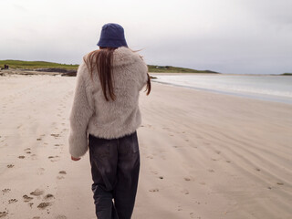 Young woman walks on sandy beach with waves crashing behind her. She wears fluffy coat and dark jeans, looking directly at camera. Her expression is calm and confident under overcast sky. Travel theme © mark_gusev