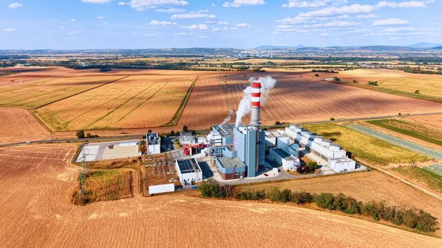 A vast grid of golden harvested fields with a factory facility under a cloudy blue sky. A large power plant or manufacturing complex surrounded by a patchwork of rural farmlands. Aerial view.