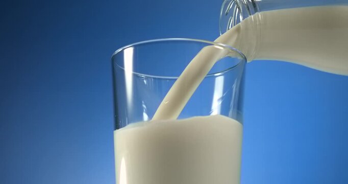Milk pouring from bottle into glass on blue background in slow motion