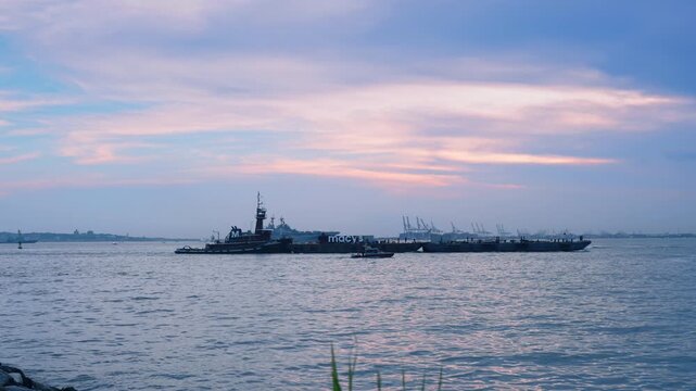 Scenic view of a body of water under a soft evening sky filled with light pink and blue clouds. A tugboat is towing a long, flat barge that has a "macy's" logo printed on its side.